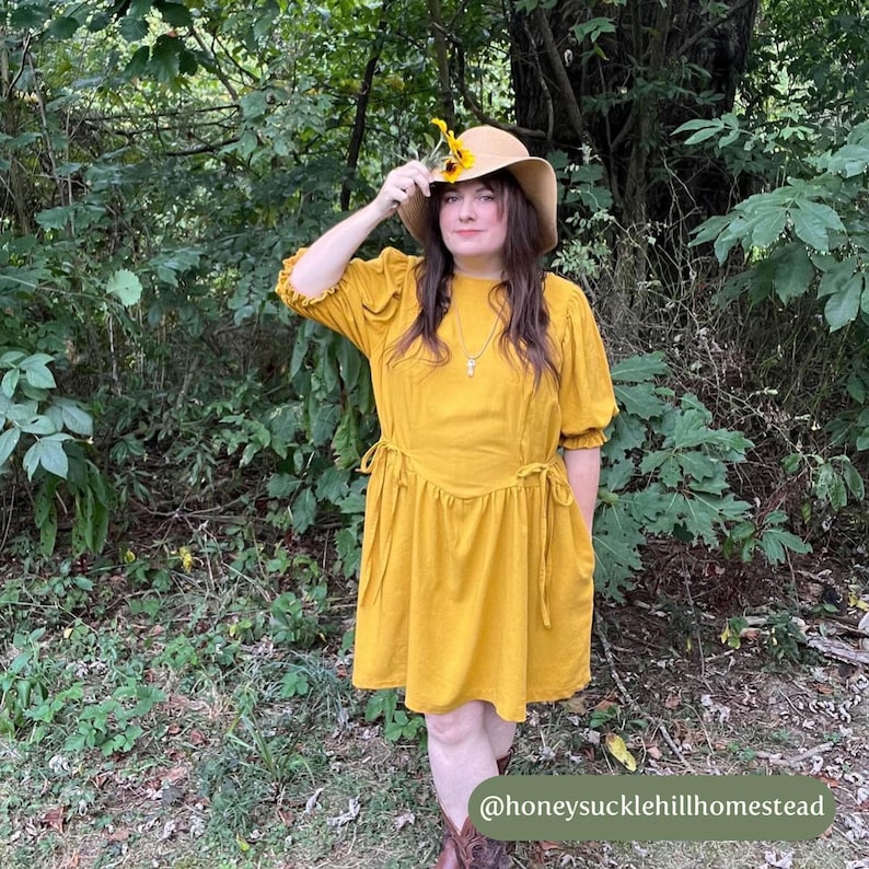May include: A woman wearing a mustard yellow dress with a cinched waist and puff sleeves. She is wearing a straw hat with a sunflower on it and brown boots. The woman is standing in a wooded area. The text "@honeysucklehillhomestead" is at the bottom of the image.