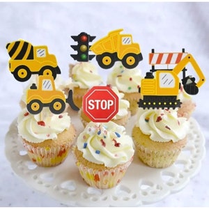 May include: Six yellow and black construction vehicle cupcake toppers with a red stop sign topper. The toppers are on cupcakes with white frosting and colorful sprinkles.