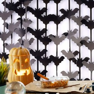 May include: A carved pumpkin with a lit candle inside sits on a table in front of a black and white bat-shaped paper garland.