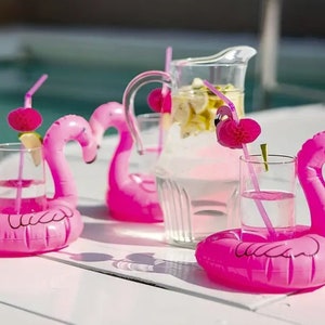 May include: Bright pink inflatable flamingo drink holders with clear glasses and pink straws. A clear pitcher with lemon slices is in the background. The scene is set near a pool, suggesting a summer party or poolside relaxation.
