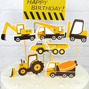 May include: A yellow cake decorated with construction vehicle themed cake toppers. The toppers include a bulldozer, a cement mixer, a dump truck, an excavator, and a crane. The cake is decorated with white pearl sprinkles and a yellow banner that reads "Happy Birthday!"