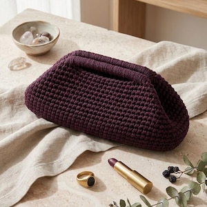 May include: A deep purple, crocheted clutch bag with a folded top, resting on a light-colored surface. A gold ring with a black stone, a gold lipstick tube, and a small bowl with stones are also present.