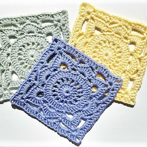 May include: Three square crochet motifs in pastel colors. One motif is light sage green, one is pale yellow, and one is periwinkle blue. Each piece features a circular, intricate, openwork pattern radiating from the center to the square edges.