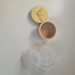 May include: A set of three beige and white silicone moulds. One mould is a round container with a lid featuring a bee design. The other two moulds are round and have four sections.