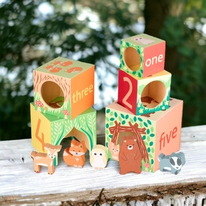 Puede incluir: Bloques de madera apilables con ilustraciones de animales y números del uno al cinco. Los bloques están pintados en tonos de verde, naranja, rojo y marrón. Los bloques están dispuestos en una torre con el número uno en la parte superior y el número cinco en la parte inferior. Hay pequeñas figuritas de animales de madera junto a los bloques.