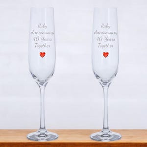 Könnte beinhalten: Zwei klare Champagnerflöten mit einem roten Herz-Anhänger an jeder. Die Flöten sind mit dem Text "Ruby Anniversary 40 Years Together" graviert.