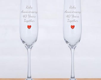 May include: Two clear champagne flutes with a red heart charm on each. The flutes are engraved with the text "Ruby Anniversary 40 Years Together".
