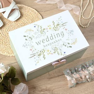 May include: A white wooden keepsake box with a light green lid. The box has a floral design with the words "wedding keepsakes" printed on the front. The box has a butterfly-shaped knob.