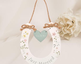 May include: White horseshoe-shaped wedding decoration with floral accents and the words "Just Married." A small, light blue heart with "You & Me" hangs in the center. The decoration is suspended by a twine loop.