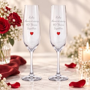 May include: Two clear glass champagne flutes featuring the engraved text Ruby Anniversary 40 Years Together and a small red heart icon. The tall, slender stems support the bowls, which are designed for sparkling wine or celebratory toasts.
