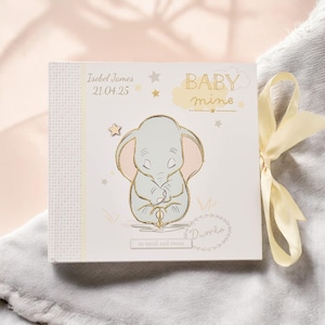 Puede incluir: Un libro de recuerdos para bebés de color blanco con una cinta amarilla. La portada presenta una ilustración de Dumbo el elefante con el texto "Baby mine" y "so small and sweet".