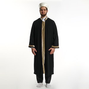 May include: A man wearing a black robe with gold trim and a white hat. The robe has a long, flowing design and is worn over a white shirt. The man is standing on a white background.