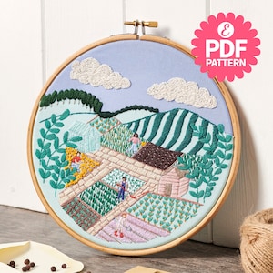 May include: Embroidered hoop art featuring a whimsical countryside scene with a blue sky, white clouds, green hills, and a small village with houses and gardens. The hoop is made of wood and has a light blue fabric background.