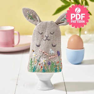 May include: A gray linen bunny egg cozy with embroidered flowers and a bee. The cozy is sitting on a white egg cup.