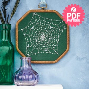 Könnte beinhalten: Eine achteckige, holzgerahmte Stickerei mit einem weißen Spinnennetz-Design mit Perlenakzenten auf grünem Hintergrund. Das Kunstwerk hängt an einer blauen Wand. Eine rosa blütenförmige Grafik mit dem Text "PDF PATTERN" befindet sich oben rechts.