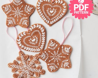 Peut inclure: Cinq décorations de Noël en feutre en forme d'étoile, de cœur et de flocon de neige. Les décorations sont brunes avec des broderies blanches. Les décorations sont suspendues à un ruban rose.