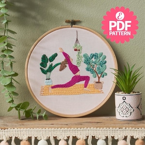 May include: Embroidered hoop art featuring a person doing yoga on a yellow mat with green plants in pots. The hoop is made of wood and has a white background.