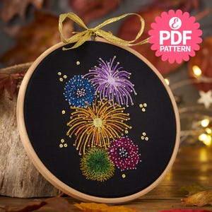 Può includere: Un telaio da ricamo nero con un colorato disegno di fuochi d'artificio. Il disegno include fili blu, viola, gialli, verdi e rosa, con accenti dorati. Il telaio è adornato con un nastro dorato e il testo "PDF PATTERN".