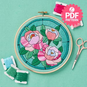 May include: A finished embroidery hoop with a stained glass design featuring three pink and white peonies with green leaves on a teal background. The hoop is surrounded by embroidery floss and scissors.
