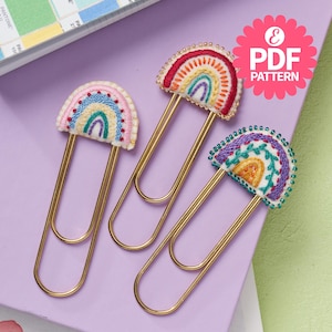 May include: Three gold paper clips with embroidered rainbow designs. Each clip features a different color scheme and design, including a rainbow with beads, a rainbow with leaves, and a rainbow with a simple design.