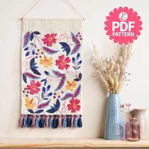 May include: A cream-coloured wall hanging featuring embroidered flowers and leaves in pink, yellow, and blue. The design includes a row of tassels at the bottom. The image also includes a pink PDF pattern logo.