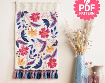 May include: A cream-colored wall hanging with embroidered flowers and leaves in pink, yellow, and blue. The design includes a row of tassels at the bottom. The image also includes a pink PDF pattern logo.