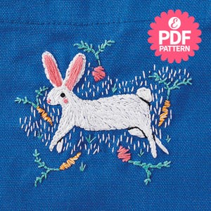 May include: White embroidered bunny rabbit with pink ears hopping through a field of green grass and orange carrots. The bunny is surrounded by small pink flowers and green leaves. The embroidery is on a blue fabric background.