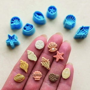 May include: A selection of blue seashell-shaped cookie cutters and miniature seashell decorations in various colours. The decorations include starfish, shells, and sand dollars, ideal for crafting or baking.