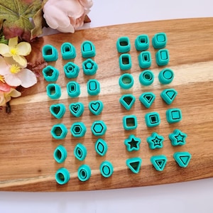 May include: A selection of teal polymer clay cutters in diverse shapes, such as hearts, stars, and geometric forms. The cutters are arranged on a wooden cutting board, suitable for crafting or baking. The shapes include circles, squares, and triangles.
