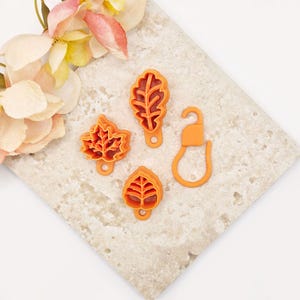May include: Three orange plastic leaf-shaped cookie cutters with intricate vein details and a small circular hole at the base of each, accompanied by a single orange plastic hook-shaped connector piece with a rounded loop at the bottom.