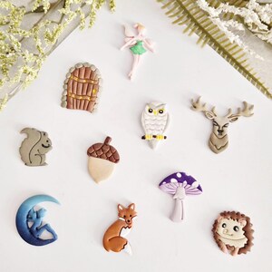 May include: A selection of charming woodland-themed embellishments. Features a fairy, owl, deer, squirrel, acorn, fox, mushroom, hedgehog, door, and a crescent moon with a figure. These decorative pieces are in various colours and designs.
