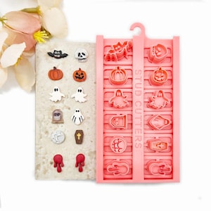 May include: A set of Halloween-themed stud earring cutters in pink plastic, featuring shapes like bats, pumpkins, ghosts, tombstones, and coffins. Beside the cutters are finished clay stud earrings in matching designs, including a skull and moon.