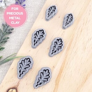 May include: Several leaf-shaped metal clay cutters are displayed. Each cutter features a grey border and a black leaf design within. The image includes the text "FOR PRECIOUS METAL CLAY" in a pink circle, suggesting their intended use.