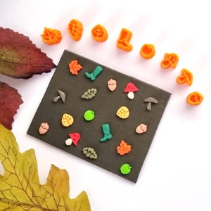 May include: A black card with various colourful polymer clay charms, including leaves, acorns, mushrooms, boots, and apples. The charms are arranged in a random pattern on the card. There are also several orange polymer clay charms in the shape of leaves, boots, and acorns around the card.