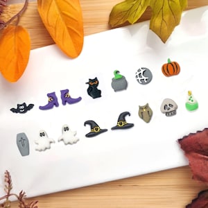 May include: A set of 14 Halloween-themed earrings featuring various spooky designs, including a black cat, a witch's hat, a ghost, a pumpkin, a cauldron, a bat, a skull, a coffin, and a pair of witch's boots. The earrings are made of polymer clay and are painted in various colours.