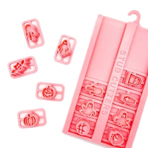 May include: A set of light pink plastic stud cutters for clay, featuring various Halloween shapes including ghosts, pumpkins, bats, a coffin, and a tombstone. The set includes a rectangular storage tray with the text STUD CUTTERS embossed on it.