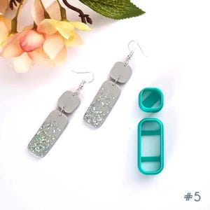 May include: A pair of grey dangle earrings with silver glitter and silver hooks. The earrings have a rectangular shape with a square top. Also shown are two teal polymer clay cutters, one square and one rectangular.