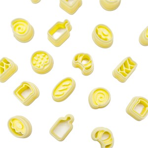 May include: A selection of yellow butter moulds in diverse shapes, including swirls, bread, and a bottle. The moulds are arranged on a white background, creating a pattern. The butter moulds are made of a light yellow material.