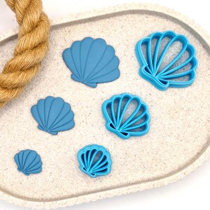May include: Five blue plastic seashell cookie cutters of varying sizes on a white sandy surface.