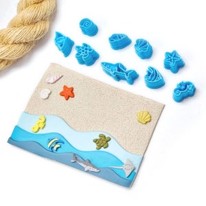 May include: A set of blue plastic cookie cutters shaped like various sea creatures, including a starfish, a turtle, a seashell, a fish, and a shark. The cutters are arranged on a white surface with a piece of brown rope in the background.