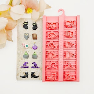 May include: A set of twelve small Halloween-themed stud earrings featuring cats, owls, books, eyeballs, potions, cauldrons, hats, and boots, displayed next to a pink plastic mould tray labelled STUD CUTTERS with matching shapes for crafting.