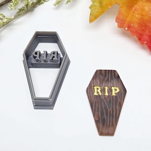 May include: A grey plastic coffin-shaped cookie cutter and a wooden-textured coffin-shaped decoration, both with the letters "RIP". Ideal for Halloween-themed baking projects.