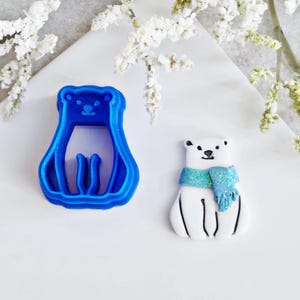May include: A blue plastic polar bear-shaped cookie cutter beside a decorated polar bear biscuit. The biscuit is white with a blue scarf and black detailing.