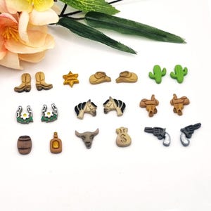 May include: A set of 16 western-themed earrings featuring various designs, including cowboy boots, horseshoes, cacti, and a cowboy hat. The earrings are made of a light brown material and have a rustic look.