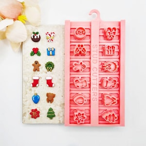 May include: A pink plastic stud cutter mould featuring twelve distinct holiday-themed shapes, including a gingerbread man, stocking, and tree. Beside the mould is a display board with twelve corresponding finished clay charms in festive colours.