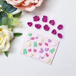 May include: A set of pink plastic cookie cutters in various shapes, including hearts, birds, bears, cupcakes and roses. The cutters are arranged on a pink card with a variety of colourful, pre-cut shapes.