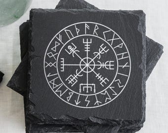 May include: A stack of square, black slate coasters. Each coaster features a white, circular Viking compass design. The coasters are made of a natural stone material and are suitable for use with hot or cold beverages.