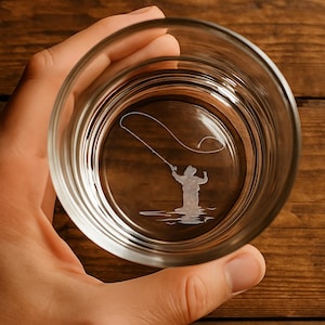 May include: A clear glass tumbler with a detailed etched design of a person fly fishing. The design is centered in the bottom of the glass. The glass is held in a hand against a wooden background.