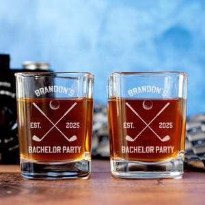 May include: Two square shot glasses filled with amber liquid. Each glass is etched with "Brandon's", a golf ball, crossed golf clubs, "EST. 2025", and "Bachelor Party". A vintage camera and patterned tie are in the background.