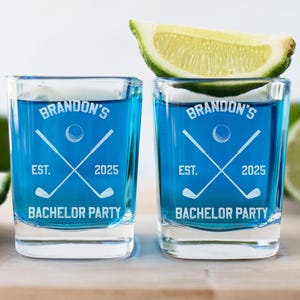 May include: Two square shot glasses filled with blue liquid, featuring a golf-themed design with the text "Brandon's Bachelor Party Est. 2025." A lime wedge rests on top of one glass. Limes are in the background.